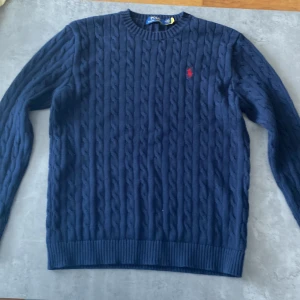 Knitted Polo Ralph Lauren tröja - Kabelstickad mörkblå tröja från Polo Ralph Lauren med klassisk rund hals och röd broderad logga på bröstet. Tröjan är långärmad och har ribbade muddar vid ärmslut och nederkant. Perfekt för dig som gillar stilren och tidlös design.