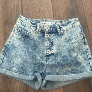 Jeansshorts - Snygga högmidjade jeansshorts från FB Sister i acid wash blått. Klassisk femficksmodell med uppvikta benslut och fyra silverfärgade knappar framtill. Perfekt för dig som gillar en retro vibe och vill sticka ut lite extra i sommar. Knappt använda, säljer även i mörkgrå, pris kan diskuteras💕