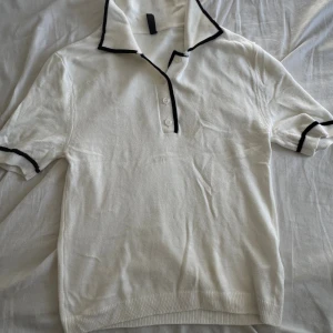 Vit pikétröja med svarta detaljer - Vit pikétröja från DeFacto med svarta kanter på krage och ärmslut. Klassisk pikékrage med tre knappar framtill. Mjuk och stickad känsla i materialet, perfekt för en clean och stilren look.