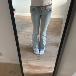 Ljusblå bootcut jeans med fickdetaljer - Snygga ljusblå jeans med bootcut-modell och coola fickor bak med lock och knapp. Jeansen har klassisk femficksdesign, låg midja och är tillverkade i mjukt denimtyg. Perfekta för dig som gillar en avslappnad men trendig look.