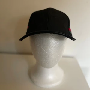 Svart Gucci keps med GG-mönster - Svart keps från Gucci med diskret GG-monogram över hela tyget och klassiska gröna och röda ränder på sidan. Justerbar rem baktill i läder med gulddetalj. Snygg och ikonisk accessoar som lyfter vilken outfit som helst.