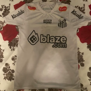 Vit Santos Neymar Jr. 10 fotbollströja - Säljer en vit Santos FC fotbollströja med Neymar Jr. och nummer 10 på ryggen. Tröjan har korta ärmar, flera sponsorloggor och klubbmärke på bröstet. Tillverkad i lätt och ventilerande material, perfekt för fotboll eller samling.