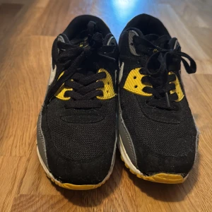 Nike Air Max 90 svart/gul sneakers - Säljer ett par Nike Air Max 90 sneakers i svart, gul och grå med vita detaljer. Skorna har klassisk Air Max-sula, mesh och skinnpaneler samt snörning. Swoosh-logga på sidorna och Air Max-detaljer i gult. Perfekta för dig som gillar streetstyle och sportig vibe.