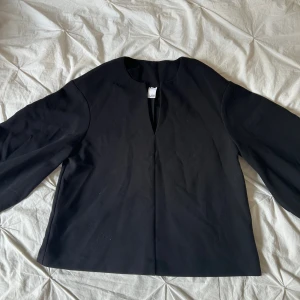 Svart blus från Visual Clothing Project - En stilren svart blus/tröja från Visual Clothing Project med lång ärm och rak passform. Blusen har en djup v-ringning framtill och är tillverkad i ett slätt tyg som ger en clean look. Perfekt för dig som gillar minimalistisk stil. Lite nopprig annars fin. 