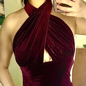 Vinröd sammet klänning - Elegant vinröd klänning i sammet med en smickrande omlott-design och hög slits. Klänningen har en halterneck-stil och är ärmlös, vilket ger en sofistikerad look.