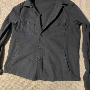 Blå overshirt från C.O.F Studio - Svart overshirt från C.O.F Studio i bomull med klassisk krage, knäppning framtill och två bröstfickor med lock och knapp. Jackan har raka fickor på sidorna och lång ärm. Perfekt för lager-på-lager och stilren look. Storlek S men passar M