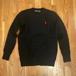 Svart kabelstickad tröja Polo Ralph Lauren - Svart kabelstickad tröja från Polo Ralph Lauren med klassisk röd broderad logga på bröstet. Tröjan har rund halsringning och långa ärmar. Perfekt för dig som gillar stilrena och tidlösa plagg med premiumkänsla.