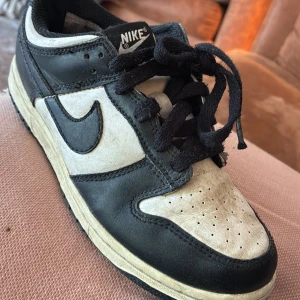 Nike Dunk Low svart/vit sneaker - Nike Dunk Low sneakers i svart och vitt skinn med klassisk Swoosh-logga på sidorna. Skon har svart snörning, perforerad tå och låg profil. Yttersulan är vit med svarta detaljer. Perfekt för dig som gillar retrostil och streetwear.