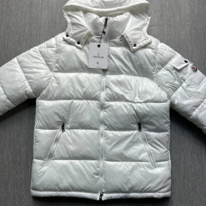 Moncler Maya - Snygg vit dunjacka från Moncler med hög krage och avtagbar huva. Jackan har två fickor med dragkedja och klassisk Moncler-logga på ärmen. Perfekt för kalla dagar och riktigt stilren look.