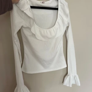 Vit blus med volangdetaljer - Supersöt vit blus med långa ärmar och volang runt den djupa ringningen samt vid ärmsluten. Perfekt för dig som gillar romantiska detaljer och en feminin look. Tillverkad i ett lätt och mjukt tyg som känns skönt mot huden.