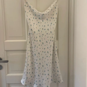 Vit blommig kortklänning med snörning - Supersöt vit kortklänning med små blå blommor och tunna axelband. Klänningen har en snygg draperad halsringning och öppen rygg med snörning som knyts i ryggen. Perfekt för sommarens alla tillfällen, lätt och flowy passform.