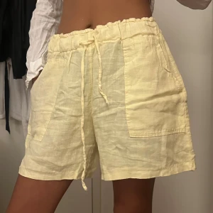 Ljusgula linneshorts med snörning - Säljer ett par ljusgula shorts i linne med hög midja och bred resår. Shortsen har snörning i midjan och stora, praktiska fickor framtill. Perfekta för varma dagar och ger en chill vibe med sin loose passform.