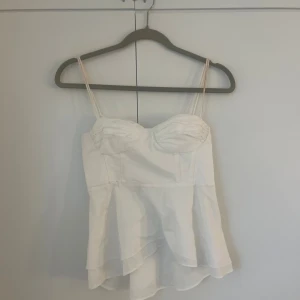 Vit linnetopp med volang - Säljer en söt vit linnetopp med tunna axelband och volang nertill. Toppen har markerade kupor och en lätt utsvängd passform som ger en romantisk vibe. Perfekt för varma dagar eller när du vill känna dig extra fin.