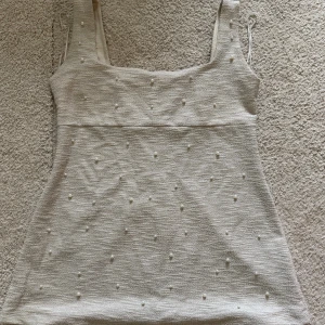 Beige kort klänning med pärlor - Supersöt beige kort klänning med fyrkantig urringning och breda axelband. Klänningen har ett stilrent pärlbroderi över hela framsidan. Använd 1 gång (på bilden).