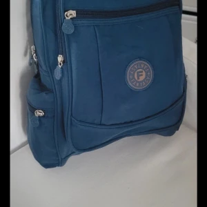  Helt ny Blå ryggsäck från Fantasy -  Helt ny Säljer en blå ryggsäck från Fantasy med flera praktiska fack och dragkedjor. Väskan har en rund logotyp framtill och är tillverkad i ett slitstarkt syntetmaterial. Perfekt för skola eller utflykter med smarta sidofickor och stilren design. 38x35x10  väldigt fint material Cresh man kan tvätta I tvättmaskin 