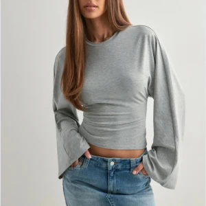 Grå topp från Nelly  - Säljer en stilren grå croppad topp med långa, vida ärmar och rund halsringning. Toppen har en tight passform över midjan och är tillverkad i mjukt bomullsmaterial. Perfekt för dig som gillar en enkel men trendig look. Använd en gång och den är slutsåld på hemsidan