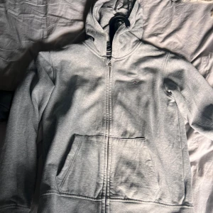 Grå hoodie från GANT med dragkedja - Säljer en stilren ljusgrå hoodie från GANT med dragkedja och huva. Tröjan har fickor framtill och en liten broderad GANT-logga på bröstet. Perfekt för chill dagar och enkel att matcha med jeans eller mjukisbyxor.