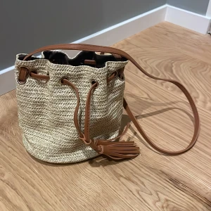 Beige axelväska med snörning - Snygg beige axelväska i flätad stil med bruna detaljer och snörning upptill. Väskan har en lång axelrem och dekorativ tofs framtill. Köpte på en marknad i Grekland❤️