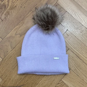 Ljuslila mössa från Steve Madden - Snygg ljuslila ribbstickad mössa från Steve Madden med stor fluffig pälsboll i brunt på toppen. Metallmärke med logga framtill. Mössan är bara provad men saknar tyvärr prislapp.