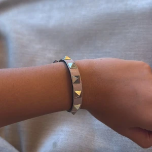 Valentino Garavani Rockstud armband - Snyggt armband från Valentino Garavani i ljusrosa skinn med ikoniska silvriga pyramidnitar. Justerbart spänne i metall och levereras med originalask, dustbag och äkthetskort. Perfekt accessoar för att lyfta din outfit med en edgy touch.