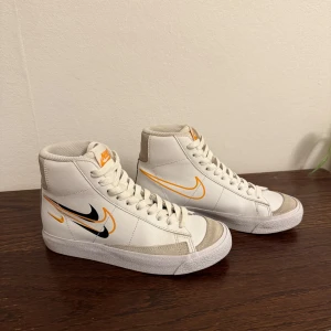 Nike Blazer Mid vita sneakers - Nike Blazer Mid sneakers i vitt skinn med orange och svart swoosh-detalj på sidorna. Klassisk hög modell med snörning och beige mockadetaljer vid tån och hälen. Snygg retrostil som sticker ut med flera swoosh-loggor och orange Nike-märke på plösen.