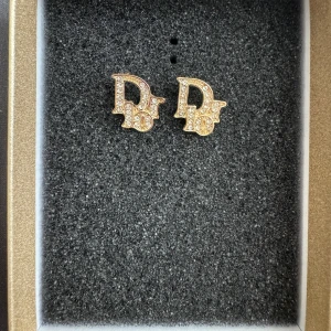 Dior örhängen med strass - Snygga örhängen från Dior i guldton med glittrande strass-stenar. Örhängena har den ikoniska 'Dior'-loggan och är perfekta för att ge din look lite extra lyx. De är små och stilrena men ändå iögonfallande tack vare alla stenar. Aldrig använda.