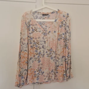 Blommig blus från KappAhl i pastell - Supersöt blus från KappAhl med blommigt mönster i ljusrosa, blått och beige. Blusen har v-ringning med knappar framtill och långa ärmar med resår vid ärmslut. Perfekt för vår och sommar med sitt lätta bomullsmaterial och luftiga passform.