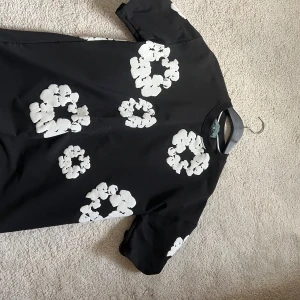 Svart T-shirt  med vita blommor Denim Tears - Svart T-shirt från Denim Tears med stora vita blommiga mönster över hela plagget. T-shirten har rund halsringning och korta ärmar. Tillverkad i mjuk bomull som känns skön mot huden. Perfekt för dig som gillar streetwear med unik twist./ Den är stor i storleken (M,L) Aldrig använt den då den är för stor på mig. 
