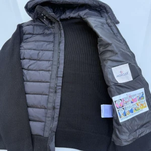 Svart Moncler cardigan med huva - Oanvänd svart cardigan från Moncler med stickade ärmar och rygg, quiltad framsida och huva. Jackan har dragkedja, innerfoder med serietidningsdetalj och praktiska fickor med tryckknapp. Perfekt för dig som vill ha en stilren och exklusiv look.