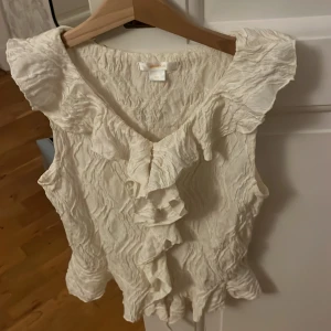 Vit volangblus från Gina Tricot - Supersöt vit blus från Gina Tricot med volangdetaljer längs v-ringningen och axlarna. Blusen är ärmlös och har ett krinklat tyg som ger en cool struktur. Perfekt för dig som gillar romantiska och trendiga plagg med extra detaljer.