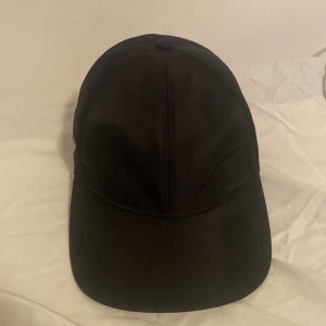 Svart keps från Prada i nylon - Snygg svart keps från Prada med klassisk böjd skärm och justerbar rem baktill. Kepsen har Pradas ikoniska metallmärke på sidan och är tillverkad i slät nylon som ger en stilren look. Perfekt för dig som gillar exklusiva accessoarer med minimalistisk vibe. Det är den gamla modellen. Priset kan diskuteras vid snabbaffär.