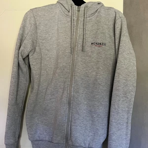 Grå hoodie från McKenzie med dragkedja - Säljer en stilren ljusgrå hoodie från McKenzie med dragkedja framtill och huva med snörning. Tröjan har ribbade muddar och diskret logga på bröstet. Perfekt för chill dagar eller när du vill ha en enkel look.