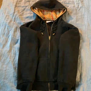 Svart Burberry hoodie med dragkedja - Svart hoodie från Burberry med klassiskt rutigt foder i huvan och guldfärgade detaljer på snörningen. Tröjan har dragkedja framtill, ribbade muddar och en skön passform. Perfekt för dig som gillar stilrena och lyxiga streetwear-plagg. Storleken M motsvara den svenska juniorstorleken 170 eller XS. 