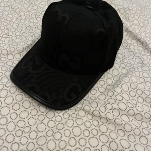 Svart keps från Gucci med GG-mönster - Snygg svart keps från Gucci med diskret GG-mönster över hela kepsen. Tillverkad i bomull och polyester med justerbar rem baktill i läderdetalj. Perfekt accessoar för en trendig och exklusiv look.