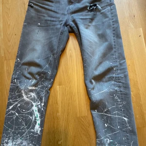 Grå jeans med färgstänk och slitningar - Grå jeans med coola vita och gröna färgstänk och slitna detaljer på fickor och lår. Raka ben och normal passform, tillverkade i klassiskt jeanstyg. Perfekta för dig som gillar en edgy och kreativ stil.