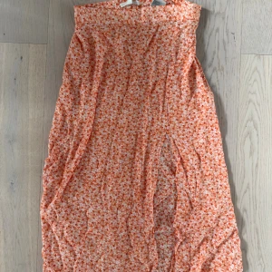 Blommig långkjol från H&M orange - Superfin långkjol från H&M i storlek 38. Kjolen har ett blommigt mönster i orange och vitt, med en rak och luftig passform. Framtill är en slits. Perfekt för dig som gillar färg och mönster! Materialet är lätt och känns skönt mot huden.