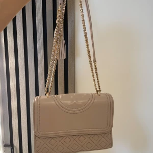 Beige/ ljusrosa axelväska från Tory Burch - Supersnygg beige axelväska från Tory Burch i skinn med guldfärgad kedja och läderrem. Tofsen är avtagbar. Har tyvärr inget kvitto kvar på den, är tyvärr lite smutsig innuti. 