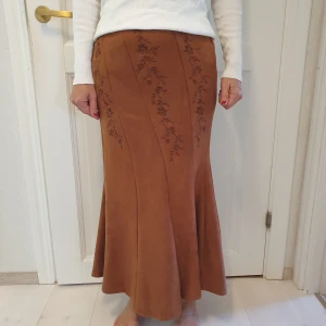 Brun långkjol med broderade blommor - Säljer en brun långkjol i mockaimitation med vackra broderade blommor längs panelerna. Kjolen har en lätt utsvängd form och känns mjuk och följsam. Perfekt för dig som gillar boho-vibbar och vill ha något unikt i garderoben.