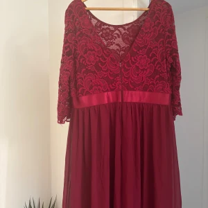 Vinröd långklänning med spetsdetaljer - Vinröd långklänning med vacker spets upptill och halvärmade ärmar. Klänningen har ett brett satinband i midjan och en flowy kjol i tunt tyg. Blommigt spetsmönster ger en romantisk vibe och den djupa v-ringningen bak gör den extra elegant.