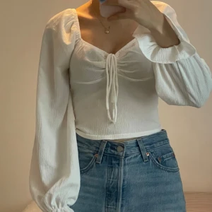 Vit blus med puffärmar och snörning - Supersöt vit blus med långa puffärmar och snygg snörning framtill. Toppen har en lätt croppad passform och är tillverkad i ett tunt, strukturerat tyg som ger en luftig känsla. Perfekt för dig som gillar romantiska och trendiga detaljer.