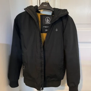 Svart vindjacka från Volcom med huva - Säljer en svart vindjacka från Volcom med huva och dragkedja. Den är väldigt bra för vinter och höst. Är i barn storlek L och passar ca max 155cm. Priset är ej satt i sten.