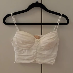 Vit korsett topp från nelly - Söt vit korsettliknande topp från nelly, med tunna  band och feminin ringning. Toppen är draperad i ryggen med dubbelt tyg, samt vaderad i bysten. Mjuk i materialet med insydd dragkedja på ena sidan.