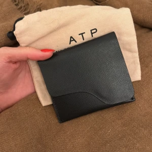 Svart plånbok i skinn från ATP Atelier - Se bilder för skick. Stilren svart plånbok från ATP Atelier i strukturerat skinn. Plånboken har en snygg vågformad kant på locket och stängs med en guldfärgad tryckknapp. Insidan har flera kortfack och plats för sedlar. Perfekt för dig som gillar minimalistisk design.