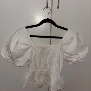 Vit puffärmad blus från BikBok XS - Supersöt vit blus från BikBok i storlek XS. Modellen har puffiga korta ärmar, smockad resår vid halsringning och midja samt volang nertill. Perfekt för en trendig och feminin look. Tillverkad i bomull för en luftig känsla.
