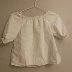 Vit blus med puffärmar och knyt - Söt vit blus med offshoulder och puffiga korta ärmar som har knyt i ändarna. Blusen har en rynkad kant upptill och är tillverkad i ett lätt bomullstyg. Perfekt för en fräsch och somrig look.