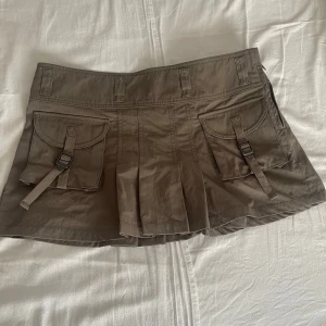 Khakigrön kortkjol från MANGO med fickor - Säljer en khakigrön kortkjol från MNG i storlek S. Kjolen har två fickor framtill med spänndetaljer och markerade sömmar. Den är lätt utställd med veck och har en cool utility-vibe. Den är helt oanvänd skick.