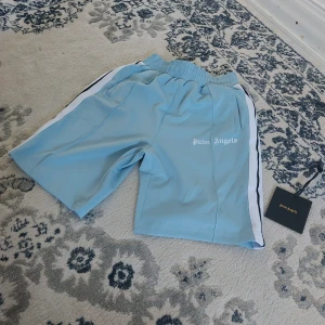 Palm Angels ljusblå shorts med revärer - Palm Angels shorts i ljusblått med vita och svarta revärer längs sidorna. Elastisk midja och logga tryckt på ena benet. Sportig vibe och skön passform, perfekta för sommaren. Materialet är polyester och shortsen har en normal passning. Palm angels lapp inkluderad åt sidan, har du frågor meddela gärna 😊