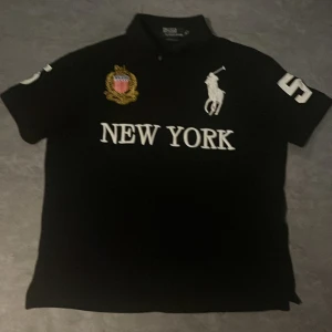 Svart Polo Ralph Lauren t-shirt New York - Svart t-shirt från Polo Ralph Lauren med vit text 'NEW YORK' och stort polospelar-tryck på bröstet. Broderad patch i guld och rött, samt siffran 5 på ärm och rygg. Klassisk krage och korta ärmar. Tillverkad i bomull.