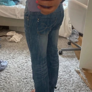 Blå bootcut jeans med broderad ficka - Snygga blå jeans med bootcut passform och låg midja. Jeansen har en cool broderad detalj på bakfickan och dubbla knappar framtill. Perfekta för dig som gillar en klassisk men trendig look.