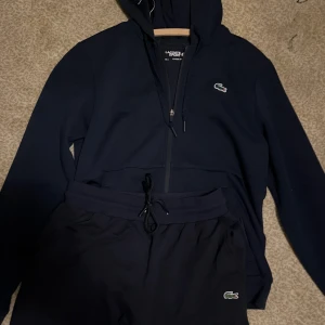 Mörkblå tracksuit från Lacoste - Snygg mörkblå tracksuit från Lacoste med klassisk krokodillogga på benet. Byxorna har resår och snörning i midjan för skön passform. Perfekta för chill eller träning, tillverkade i mjukt bomullsmaterial.